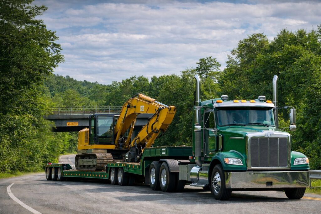 Green semi truck hauling a double drop trailer with a yellow excavator on a winding road, showing a double drop deck trailer setup for heavy equipment and taller loads.