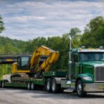 Green semi truck hauling a double drop trailer with a yellow excavator on a winding road, showing a double drop deck trailer setup for heavy equipment and taller loads.