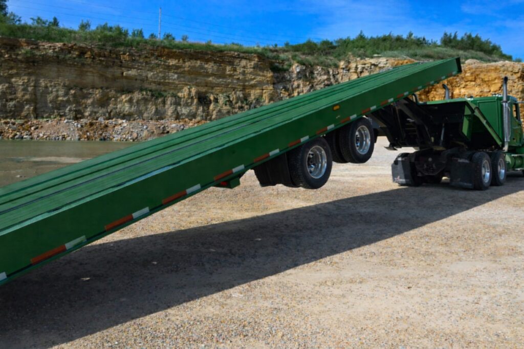 Green semi truck with a green sliding axle tilt deck trailer raised into a loading ramp in a gravel quarry, showing sliding axle trailer operation and adjustable axle position.