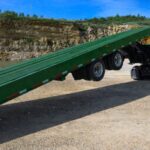 Green semi truck with a green sliding axle tilt deck trailer raised into a loading ramp in a gravel quarry, showing sliding axle trailer operation and adjustable axle position.