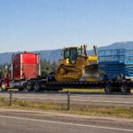 Red semi truck hauling equipment on a step deck trailer along a highway with mountains in the background
