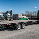 Green semi truck with a heavy-duty flatbed trailer parked in an industrial yard with construction equipment and materials nearby, illustrating flatbed trailer buying considerations for heavy loads.