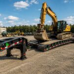 Green semi truck with RGN lowboy trailer in an industrial equipment yard, showing removable gooseneck design, low deck height, axles, tires, and heavy machinery for long-haul heavy loads.