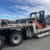 view.jpg Side view of a 2023 Extreme step deck trailer carrying a forklift, with loading ramps stored beneath the lower deck