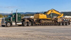 Lowboy gooseneck trailer carrying a tracked excavator on its low deck, attached to a green semi truck.