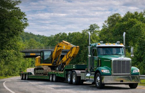 Green semi truck hauling a double drop trailer with a yellow excavator on a winding road, showing a double drop deck trailer setup for heavy equipment and taller loads.