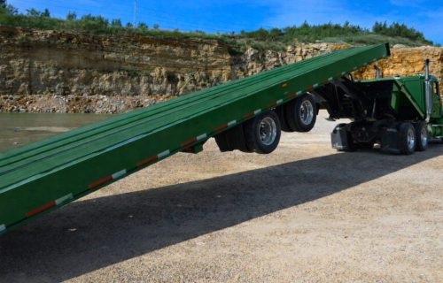 Green semi truck with a green sliding axle tilt deck trailer raised into a loading ramp in a gravel quarry, showing sliding axle trailer operation and adjustable axle position.